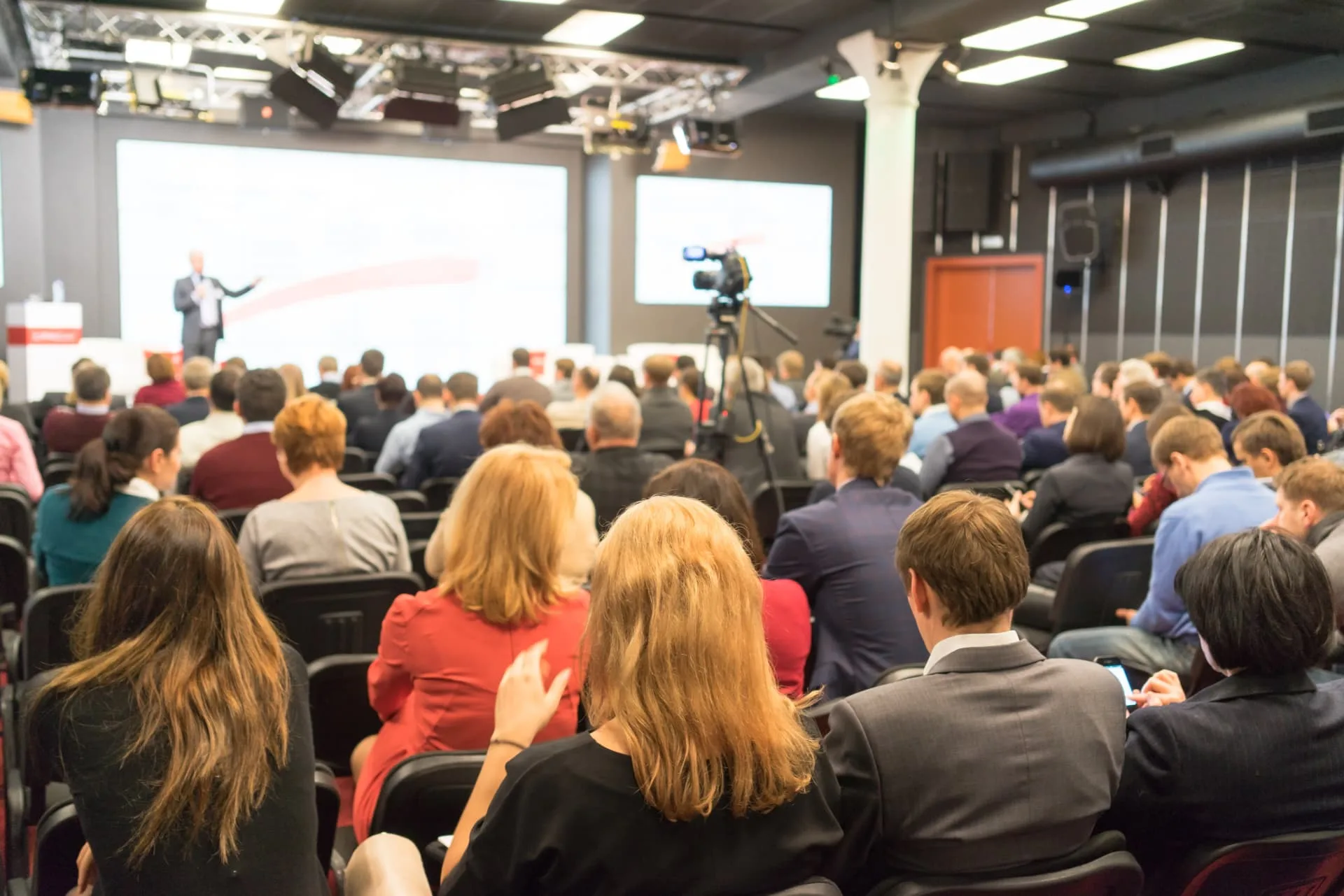 Public attentif lors d'une conférence d'entreprise, avec un intervenant sur scène et des écrans de présentation, organisé avec MADCRM